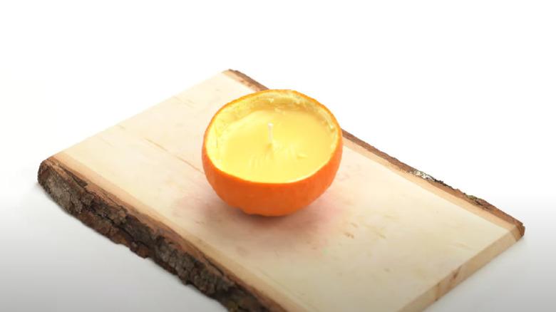 A candle made from an orange peel rind sitting on a wooden tray.