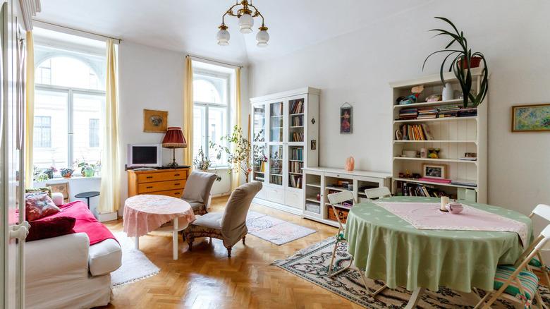 A living room with white walls, herringbone wooden floors, tables and chairs, and butter yellow curtains.