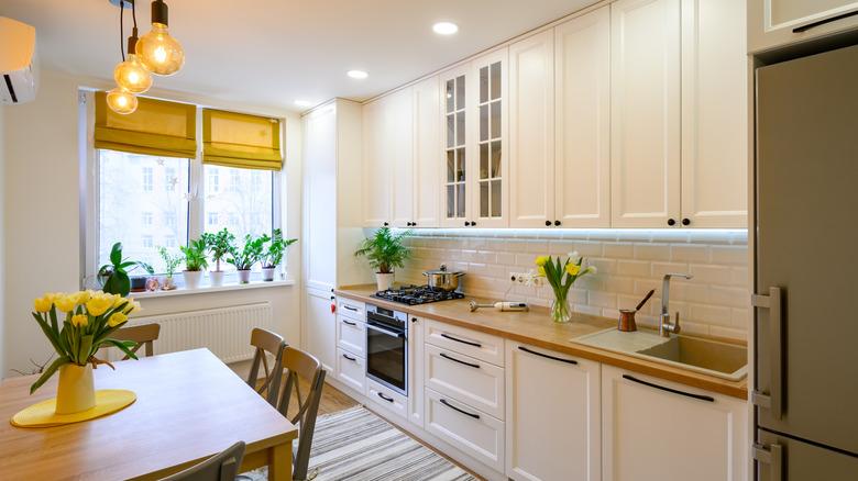 A small, galley kitchen with white kitchens and walls, as well as wooden accents and goldenrod window treatments.