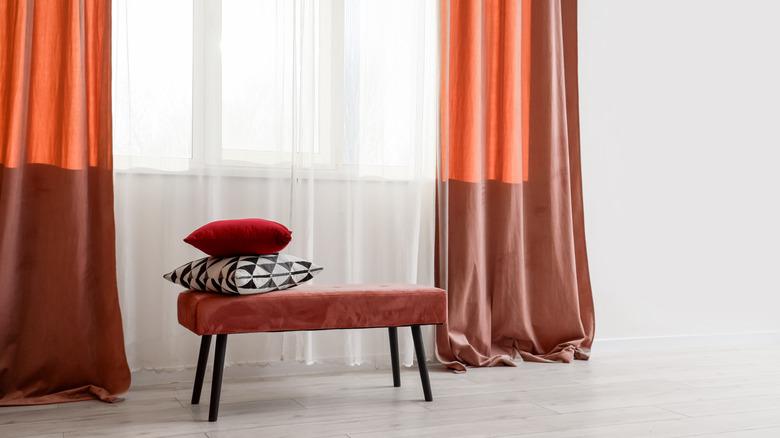 Velvet terracotta colored curtains against white walls, with a matching bench sitting on the floor.