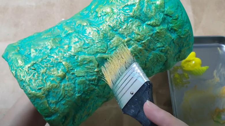 A hand painting a vase with egg carton decoupage covering it.