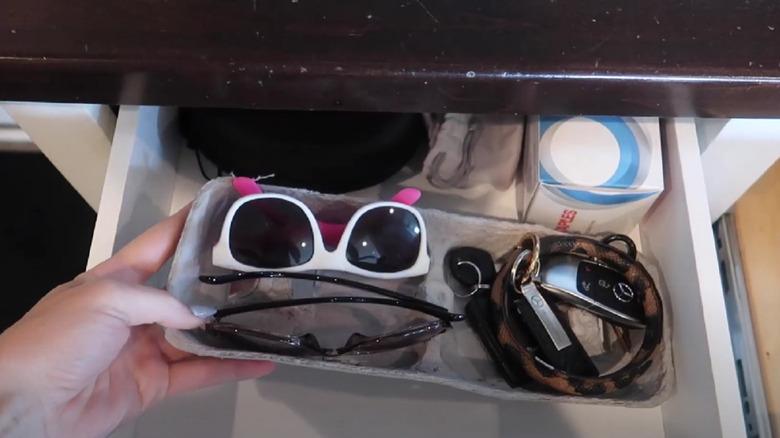 The top of an egg carton filled with sunglasses, keys, and other household items in a drawer.
