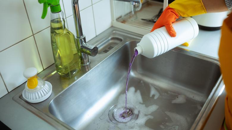 Gloved hands pouring drain cleaner down a kitchen sink.