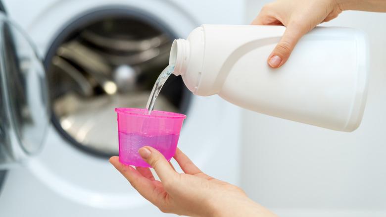 Hands pouring laundry detergent from a bottle into a measuring cup in front of an open washer.