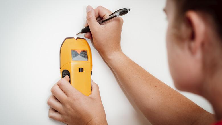 A young women holds a stud finder against a wall and uses a pen to mark the location of the building's framing stud
