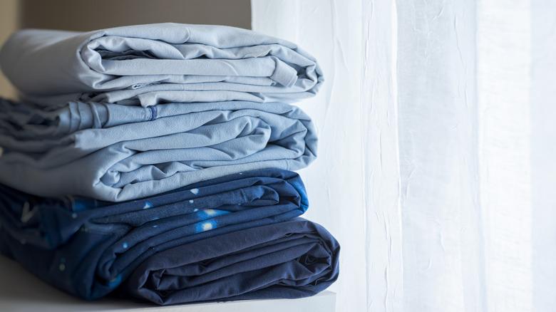 pile of folded blue bed sheets next to a sheer curtain