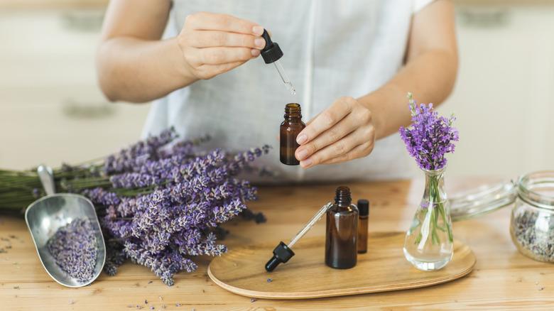a woman adding lavender essential oil to a dropper bottle, with a bundle of fresh lavender on the countertop nearby