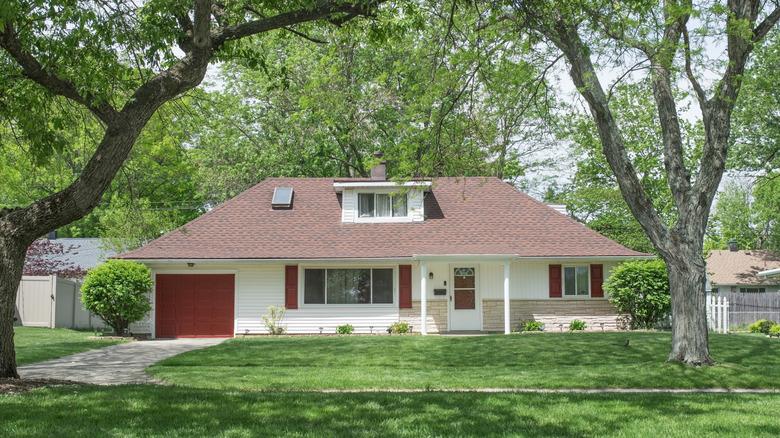 A rambler style home with a red garage, front door, and shutters, sitting among mature trees.