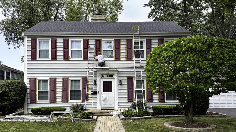 A man on a platform painting the second floor shutters of a home.