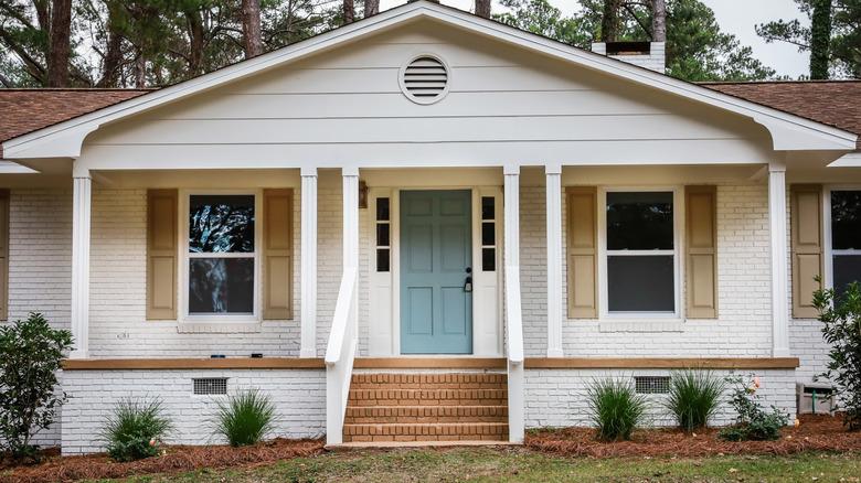 A white rambler, brick home with a blue door and khaki window shutters.