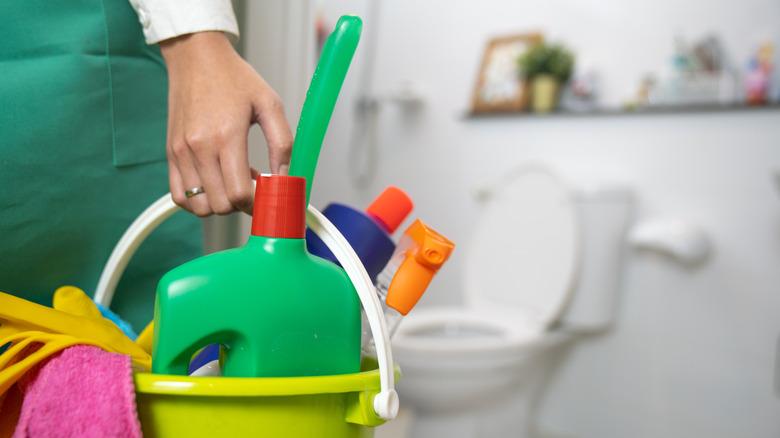 A woman holding a bucket full of cleaning supplies in a bathroom.